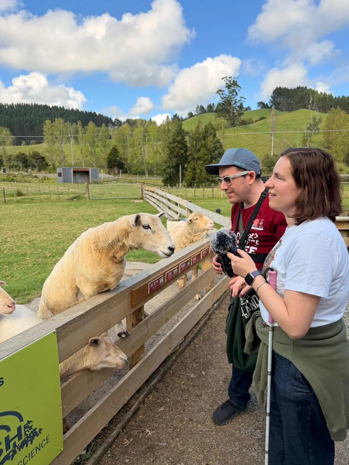 A man holds an audio recorder with a foam windscreen beside a woman holding a white cane, both standing at a wooden farm fence with sheep leaning over the rail.