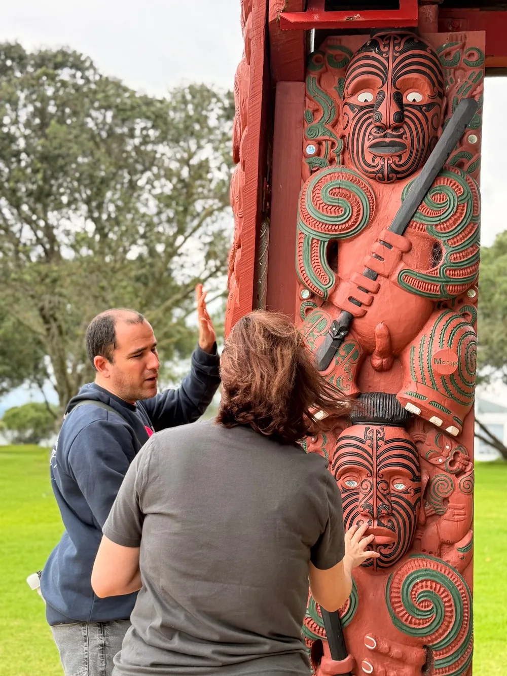 A woman rests her hand on a carved Māori figure while a companion beside her gestures as he describes it.