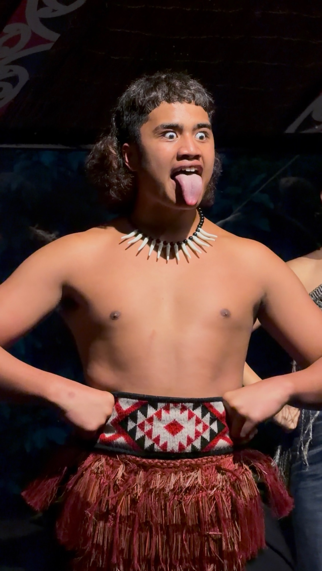 Joven maorí realizando un haka en una presentación cultural, con collar tradicional de hueso y falda piupiu, sacando la lengua en un gesto de desafío ceremonial