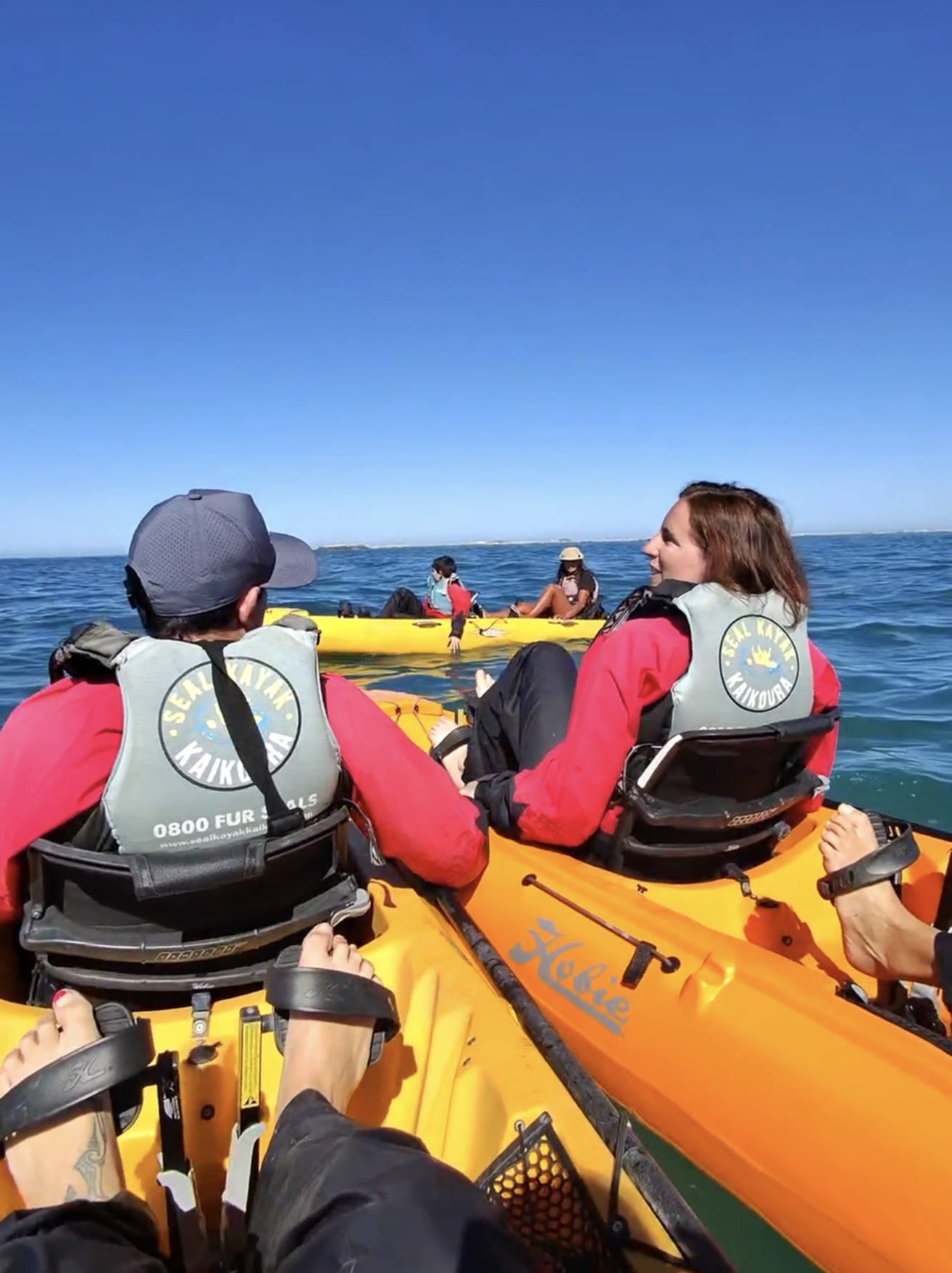 Dos viajeras sentadas en un kayak amarillo en las aguas de Kaikōura, con chalecos de Seal Kayak Kaikoura, mirando hacia otro kayak con más viajeros bajo un cielo despejado