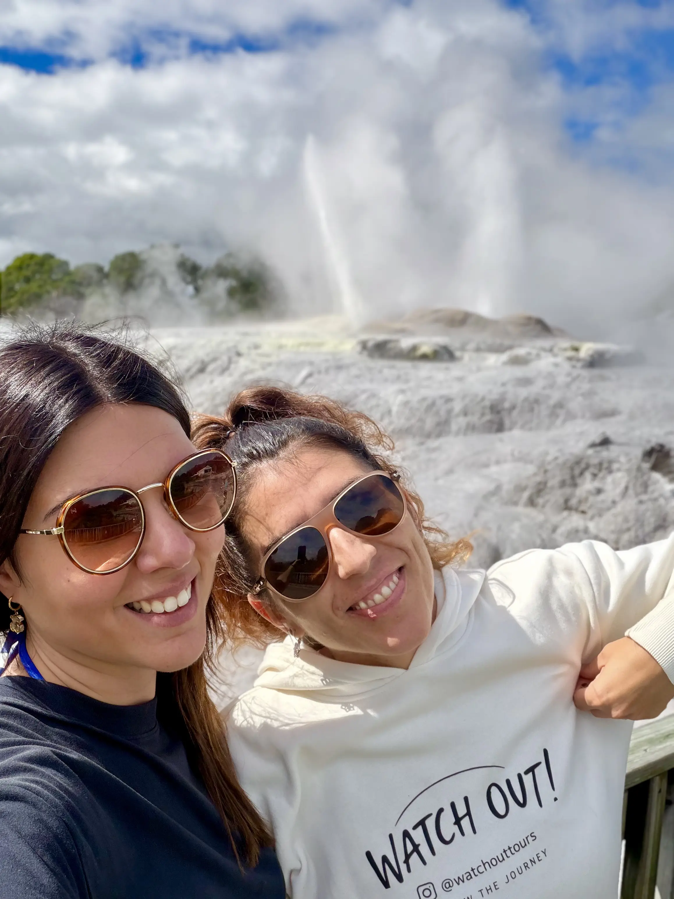 Sylvie y Moni, guías de Watchout Tours, sonriendo frente a un géiser humeante en Rotorua